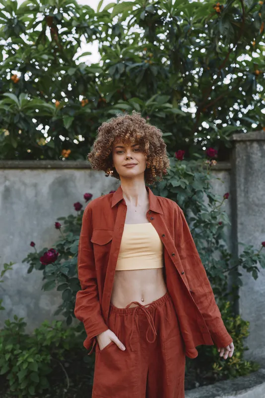 A stylish woman outdoors in a rust suit and crop top stands in a garden, smiling with relaxed confidence. Lifestyle fashion portrait in natural light and greenery