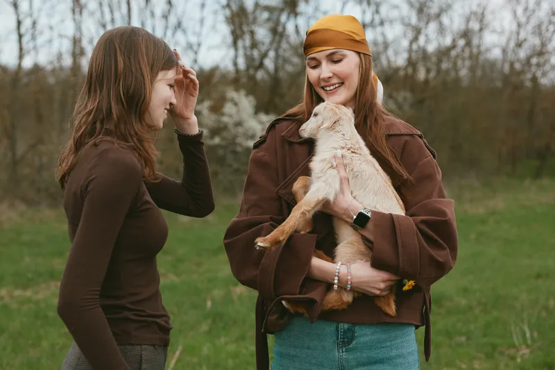 Two young women enjoy a casual outdoor moment as one holds a small goat while they smile and share a relaxed lifestyle scene in cool autumn clothing.
