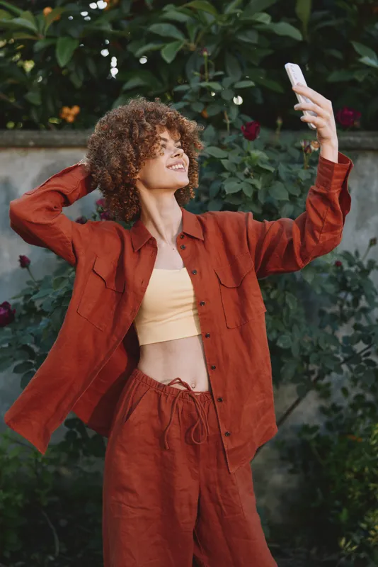 Cheerful woman outdoors, selfie in warm sunlight, rust outfit, casual lifestyle, relaxed pose, natural colors, vibrant greenery background, joyful moment