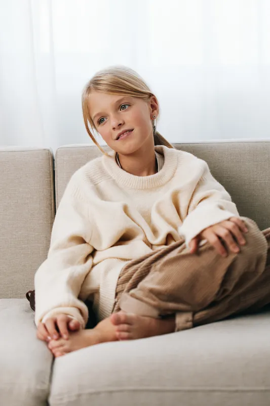 A young girl sits on a light sofa wearing a cozy cream knit sweater and soft brown pants, gazing thoughtfully off camera in a calm home setting.