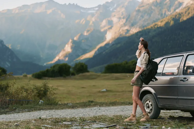 Young woman with a backpack stands beside a car on a mountain road enjoying the scenic view of the valley and peaks during a hiking trip.