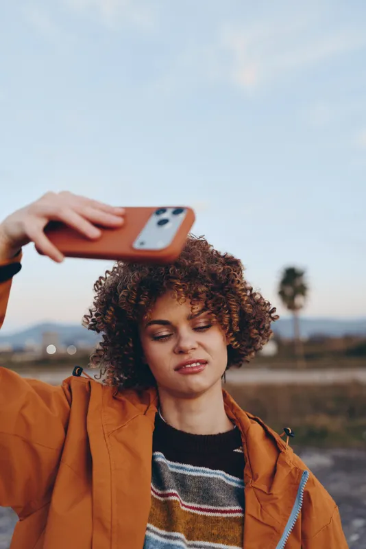 Woman lifestyle portrait outdoors at sunset, curly hair, orange jacket, smiling while holding smartphone for a selfie, hand on phone, soft sky background, relaxed travel mood
