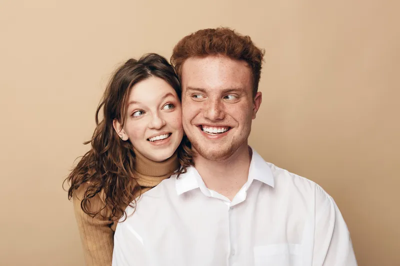 Smiling young couple with red hair and brown hair posing together on beige background. They look happy and affectionate, showing close relationship and positive emotions in casual clothing.
