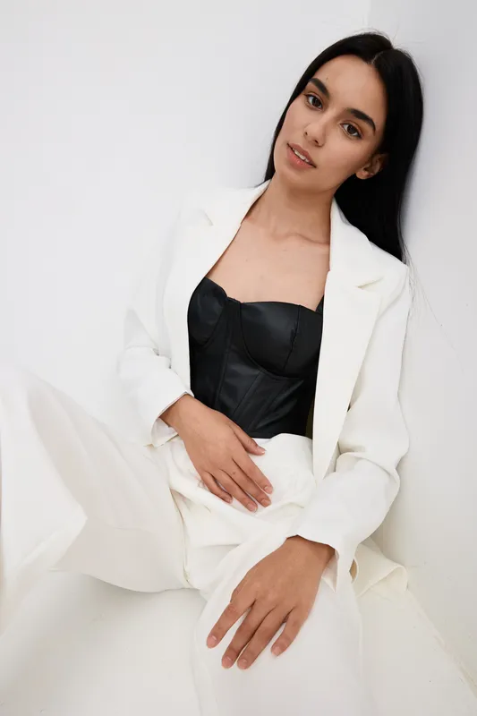 woman in white suit with black corset sitting relaxed against white background, stylish clothing, youthful, minimalistic fashion portrait