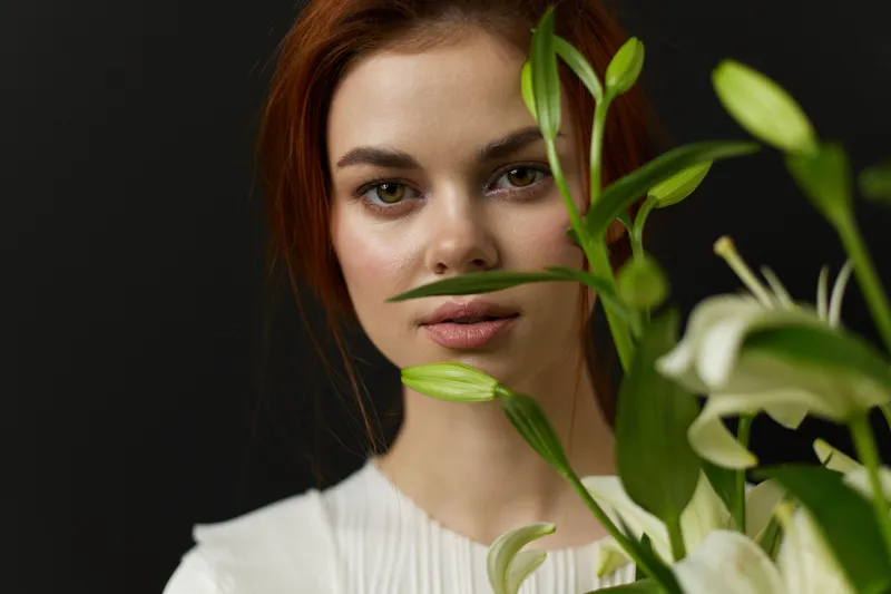 Portrait of a young woman with long red hair holding white lilies, showcasing beauty and tranquility against a dark background