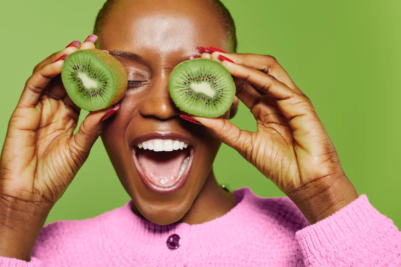 Happy dark skinned African woman in pink sweater holds two kiwi halves in front of eyes, smiling with mouth open on green background. Concept of healthy lifestyle and fresh fruit.