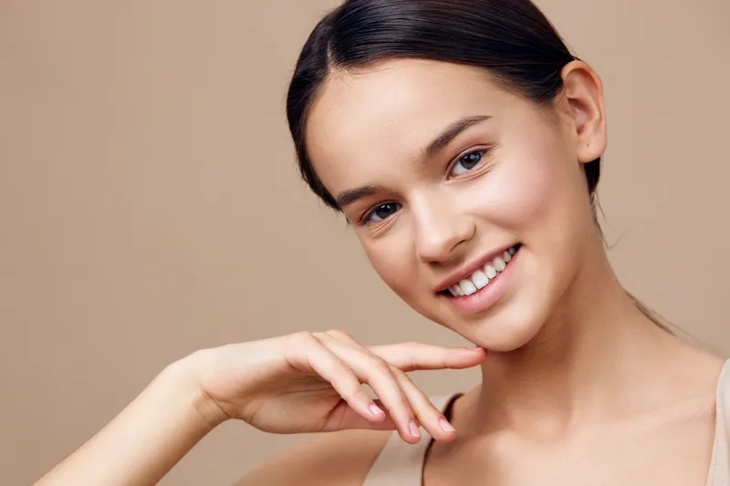 Smiling young woman with natural makeup and smooth skin against a soft beige background, showcasing beauty and confidence