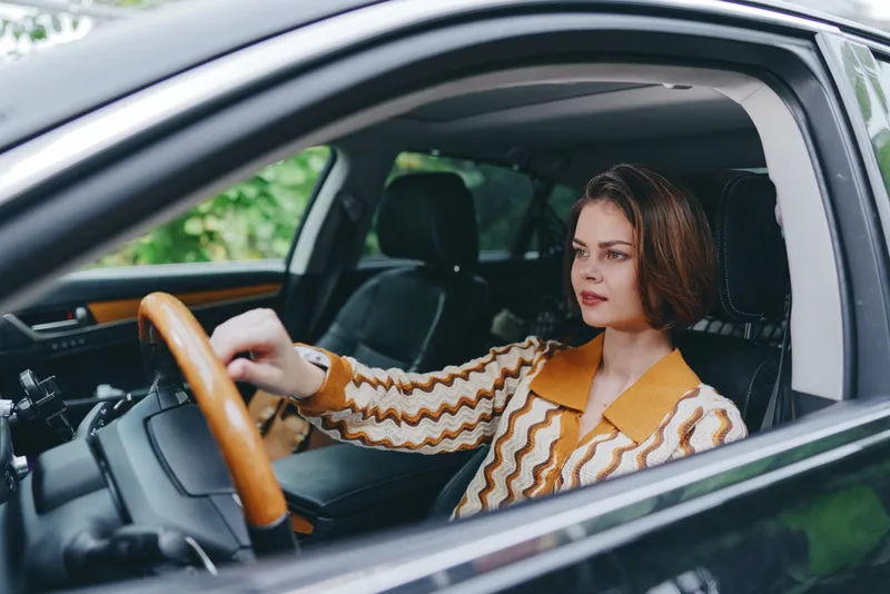 Portrait of a woman driving a car with both hands on the wheel, focused gaze, stylish outerwear, and a modern car interior framing.