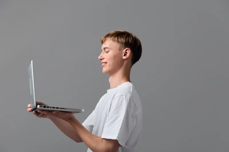 Laptop held by a smiling young man in a casual whiteshirt, shown in side view against a neutral backdrop, illustrating light workflow, digital learning, remote work, and focused engagement with