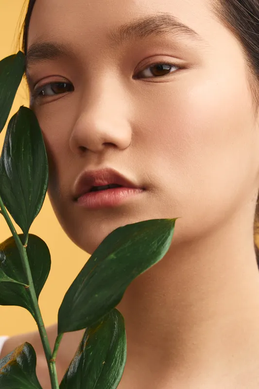 Asian woman gazes softly at the camera, framed by lush green leaves, showcasing natural beauty and calm confidence in a minimal studio setting, with warm lighting and a clean, focused portrait.