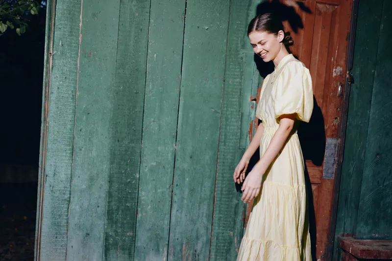 Casual portrait of a woman in a light dress walking beside a weathered doorway in an outdoor setting, with soft daylight and a vintage ambiance.