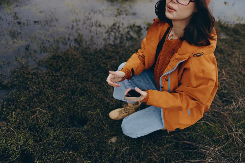 Woman lifestyle outdoors in an autumn field, orange jacket and casual jeans, sitting on dry grass, holding a smartphone, relaxed smile, warm soft sunlight, natural colors