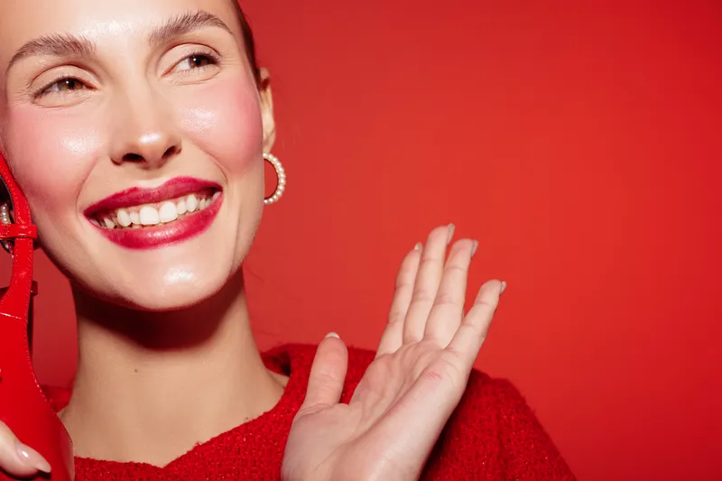Smiling woman with red lipstick and earrings waves hand on red background. Close-up portrait with bright makeup and joyful expression. Concept of happiness and beauty.