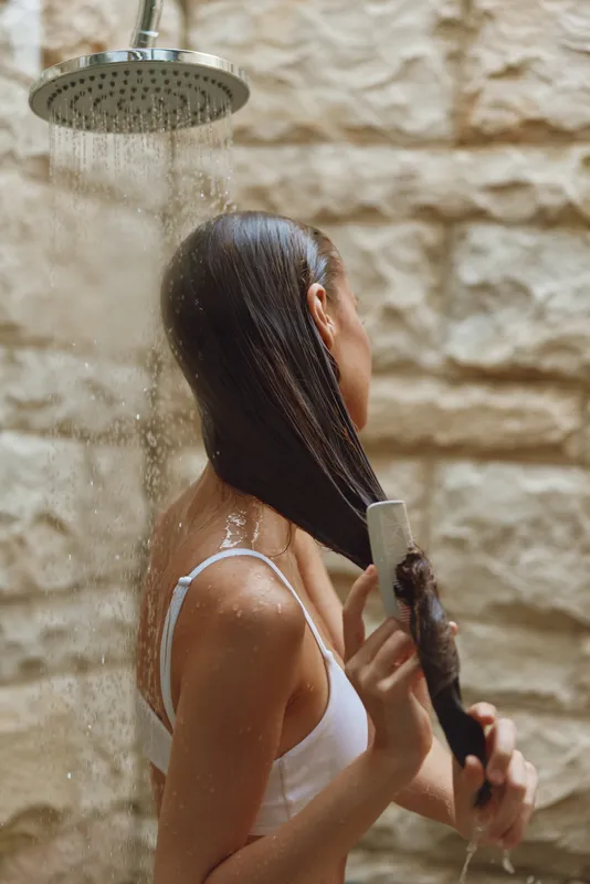 Woman washing long wet hair under shower with stone wall background, wearing white top, natural light creating a calm and refreshing atmosphere.