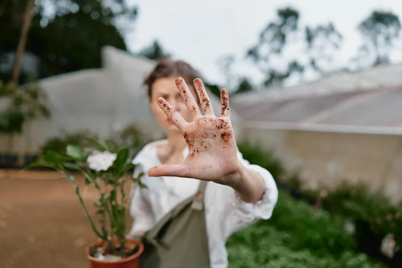 Woman with dirty hands showing a stop gesture while caring for plants, expressing joy and connection with nature in a lush garden setting