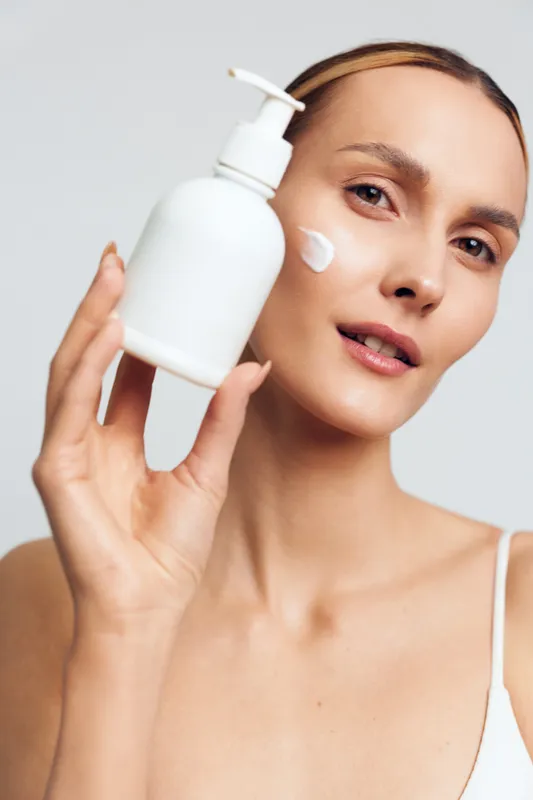 Woman applying skincare lotion, beauty face care routine, holding pump bottle by cheek, showing hydration and cosmetic care on clean white background, close up portrait with smooth skin and natural