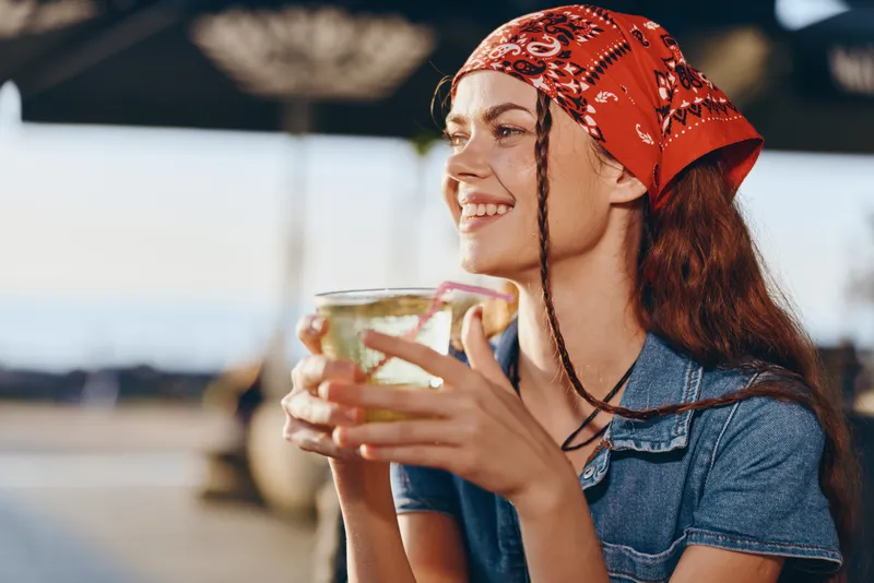Lifestyle woman wearing modern boho-western denim street style outfit with a red bandana, enjoying a drink outdoors in warm film color tones and natural light.