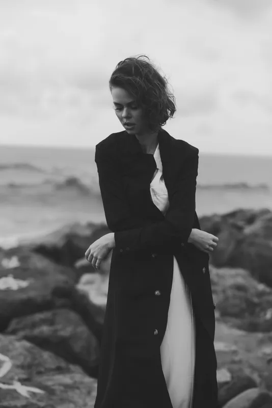 Woman in a long coat stands on a rocky coast, a monochrome scene with thoughtful mood, wind in hair, and a moody sea backdrop.