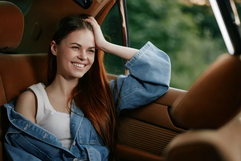 Happy young woman with long hair enjoying a road trip in a vintage car, wearing a denim jacket and smiling, reflecting carefree summer vibes in natural light.
