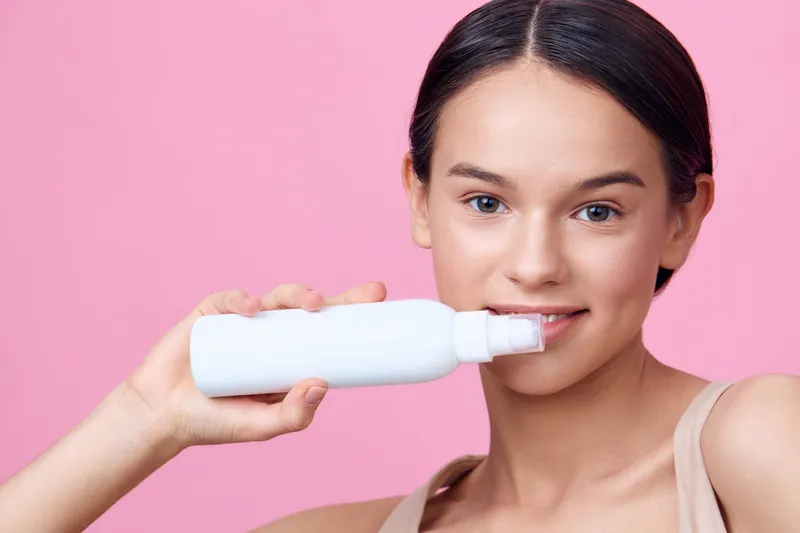 Young woman holding a white skincare bottle against a pink background, showcasing a fresh and natural beauty routine with a joyful expression