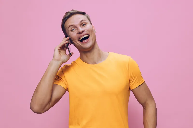 Cheerful smiling woman with short hair wearing a bright yellow t-shirt, holding a phone to her ear, looking happy and engaged, isolated on pink background. People lifestyle concept.