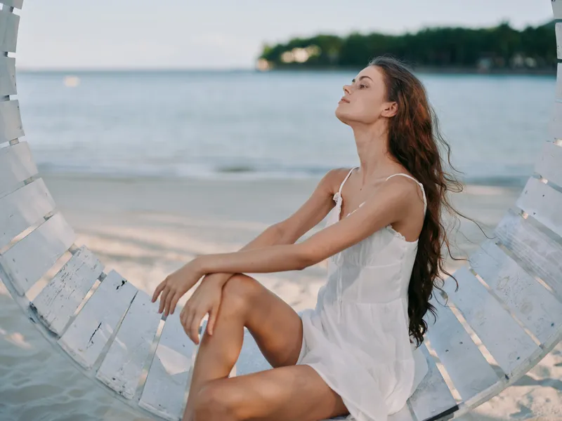 A serene woman sits on a circular swing by the seaside in a white dress, eyes closed, savoring the breeze and soft light as waves lap the shore.