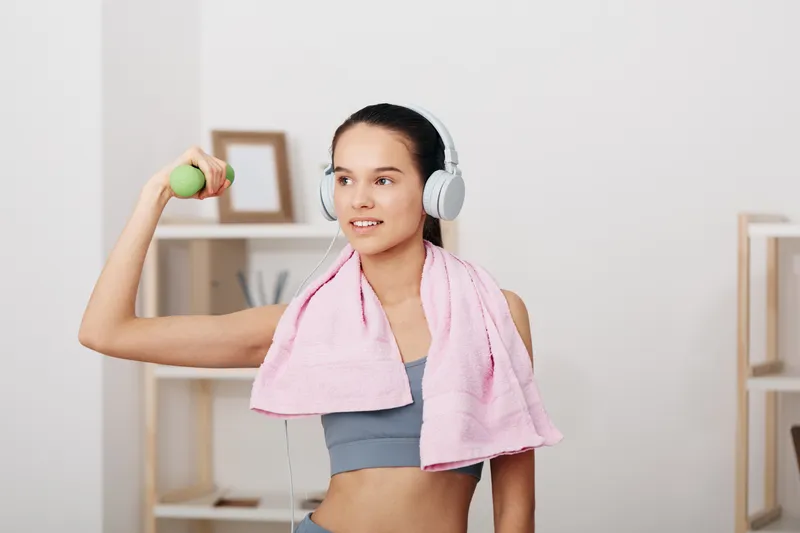 Fitness focused woman in a home gym, lifting a small dumbbell with one arm, towel draped around neck, wearing headphones and workout top, bright room, motivating routine.