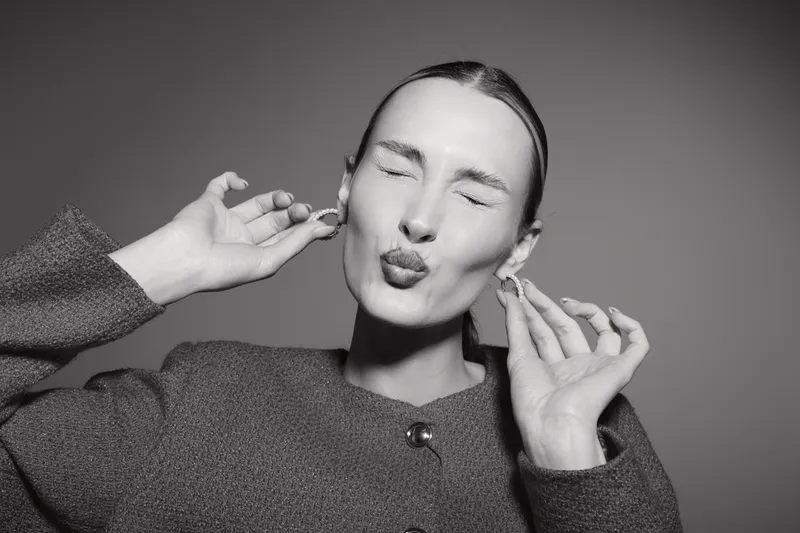 Young white woman with pale skin and slicked hair makes playful face with closed eyes and puckered lips on grey background.