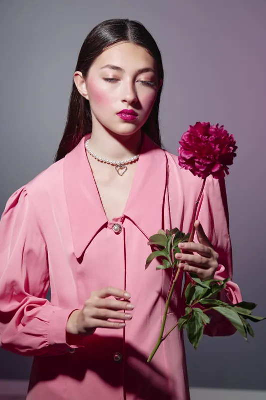 Young woman in fashionable pink outfit holding a red peony. She wears delicate jewelry and has sophisticated makeup. Studio beauty portrait with creative lighting, fashion concept.