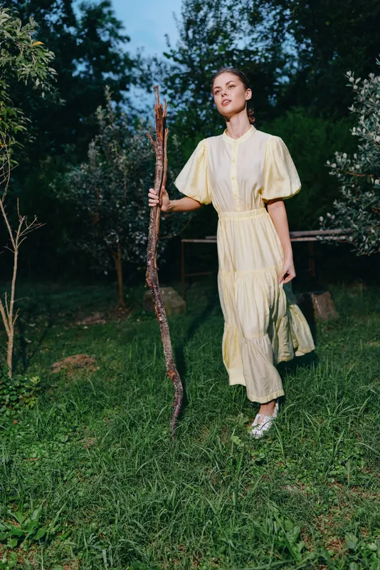 A woman in a pale yellow dress walks outdoors in a lush garden, holding a wooden staff, with calm expression and soft natural light, surrounding greenery creating a tranquil serene atmosphere