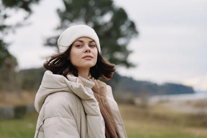 Winter scene with a woman in a warm coat and beanie standing outdoors by a lake, wind in her hair, muted tones and a tranquil natural backdrop.
