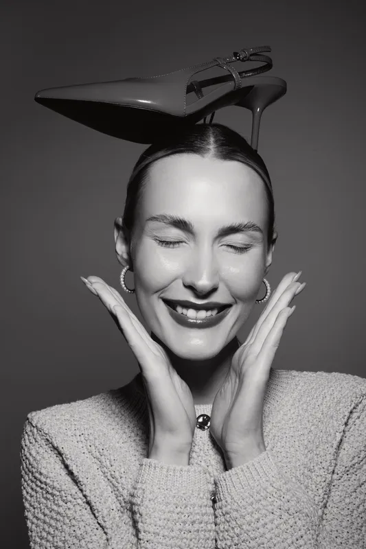 Happy Caucasian woman with fair skin and closed eyes smiles while balancing a high heel shoe on her head in studio. Concept of fashion, joy, and playful mood.