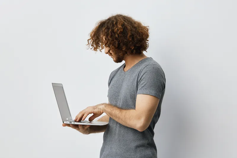 Young man with curly hair using a laptop, showcasing tech savvy attitude in a minimalist setting, ideal for remote work concepts and digital lifestyle