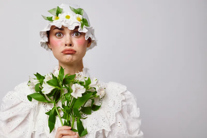 A whimsical young woman in a flower themed outfit expressing surprise while holding fresh flowers against a minimalistic white background