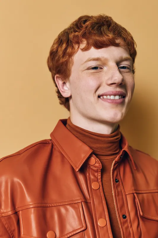 Young man with red hair wearing burnt orange leather jacket and turtleneck on beige background. Smiling face closeup with casual style and confident expression indoors portrait.