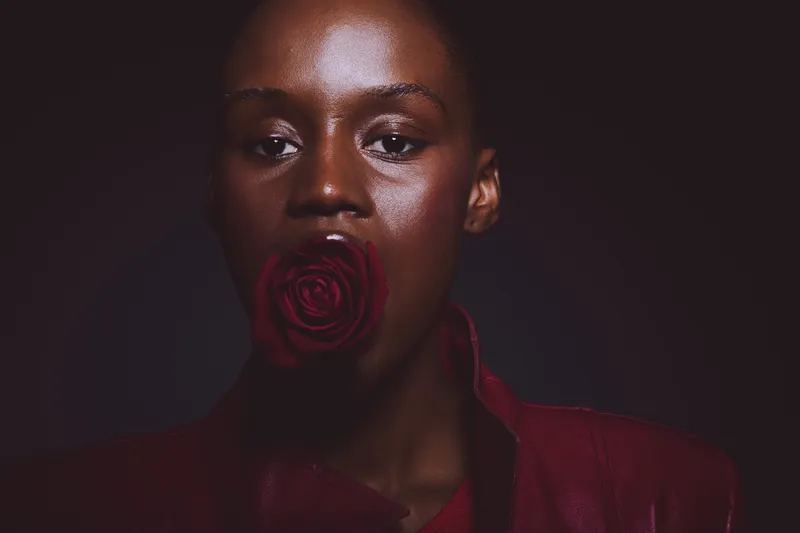 Close-up portrait of a dark skinned African woman with shiny skin holding a red rose in her mouth. The image features dramatic lighting and a dark background, emphasizing beauty and elegance.