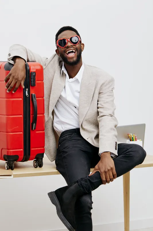 Joyful young man with dark skin in beige blazer and trendy sunglasses sitting on table with bright red suitcase isolated on white background, cheerful expression, casual style, lifestyle concept.