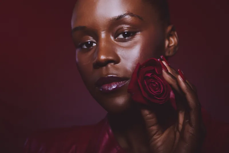 Close-up portrait of a dark skinned African woman holding a red rose near her face. The image emphasizes natural beauty, elegance, and floral aesthetics on a dark red background.