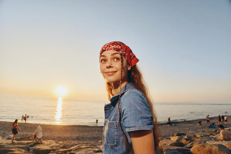 Lifestyle woman in modern boho-western denim street style smiling at sunset on the beach with warm film color tones and relaxed summer mood.