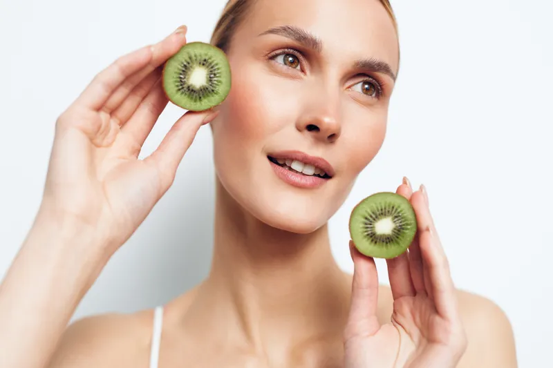 Young woman with brunette hair holds two halves of a kiwi fruit near her face while looking away with a gentle smile on a white background.