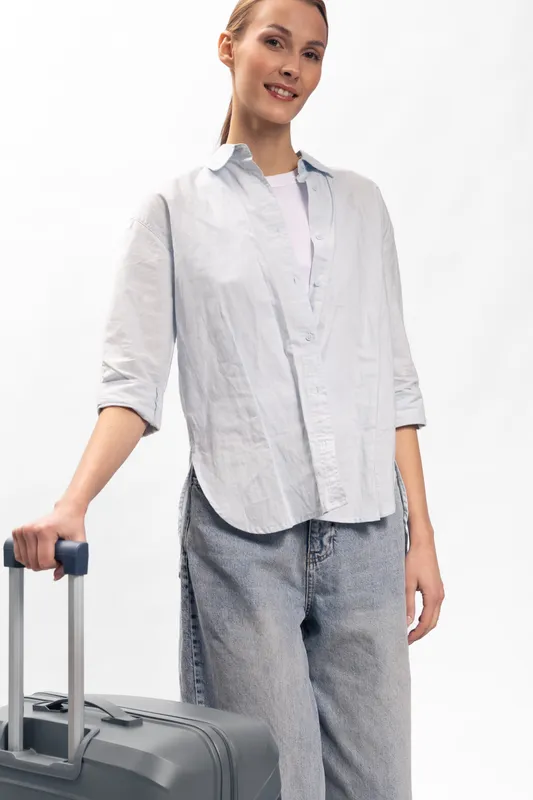 Young woman in casual clothes standing with suitcase handle, smiling and looking at camera. Travel concept with luggage, casual outfit, and positive mood on white background.