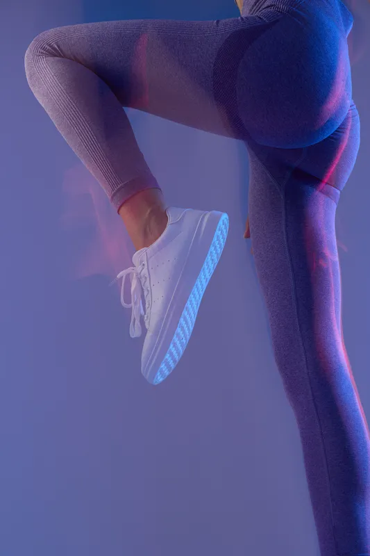 woman sport fitness wearing purple leggings and white sneakers, performing high knee exercise with dynamic lighting, showcasing active lifestyle and strength.