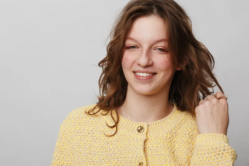 Smiling young woman with wavy brown hair wearing a yellow knitted sweater on a light background. Casual portrait with natural expression and soft lighting for lifestyle concept.