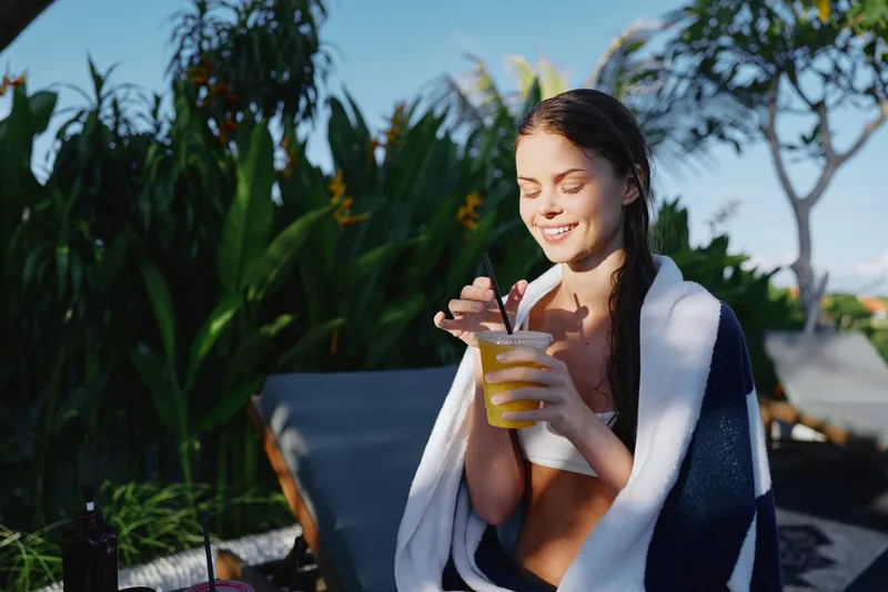 Woman enjoys a refreshing drink outdoors by the pool, wrapped in a towel, smiling in bright sunshine during a relaxed summer moment, and clear water nearby while birds sing softly.