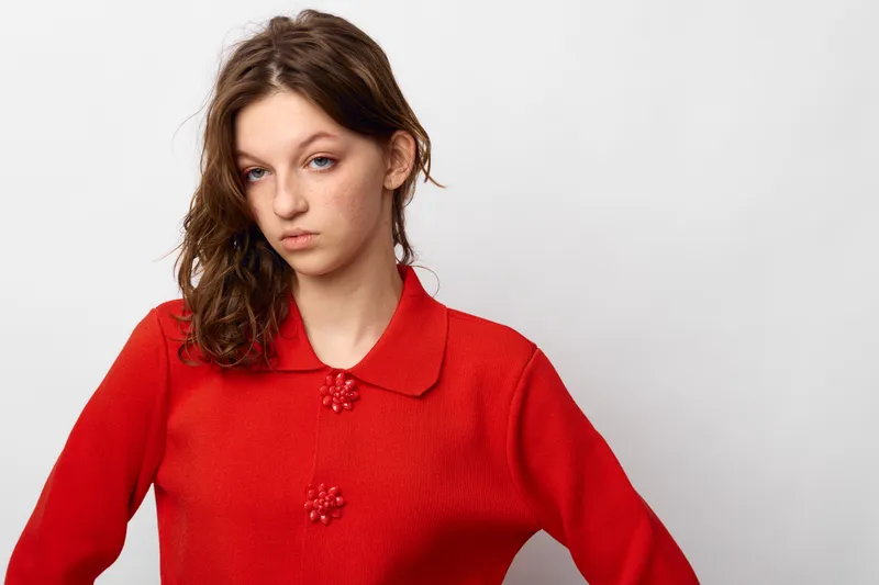 Young woman with wavy brown hair wearing a red sweater looks serious and thoughtful against a plain white background. Concept of casual fashion and natural beauty.