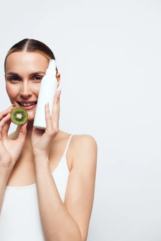 Young woman holding kiwi slice and white cosmetic bottle, clear skin, natural beauty, skincare routine, fresh fruit, minimalistic background, healthy lifestyle