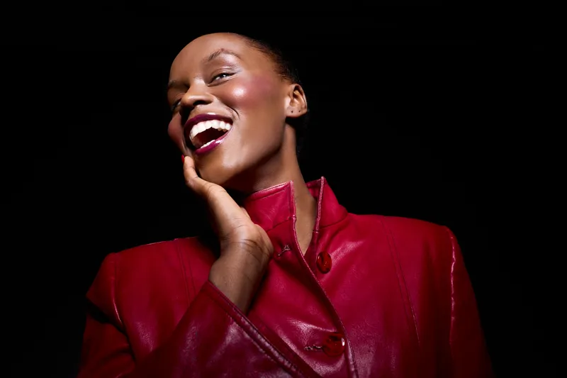 Happy African American woman with dark skin laughing and touching face, wearing shiny red jacket. Studio portrait on black background. Concept of joy, confidence, fashion, and beauty.