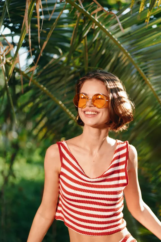 A cheerful young woman wearing a striped cropped top and sunglasses smiles as she enjoys a sunny day outdoors, radiating warmth, energy, and carefree summer vibes in a bright, lively setting.