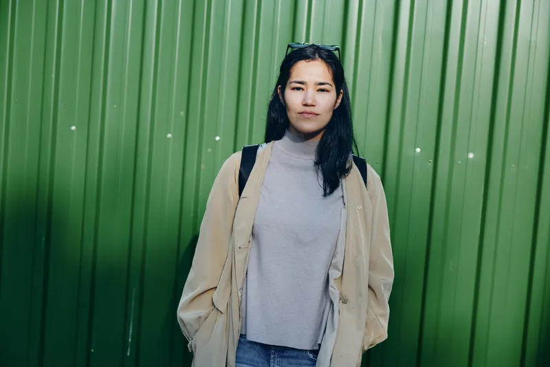 Woman wearing a beige coat and gray turtleneck stands before a green wall, casual street fashion, outdoor urban style, confident pose ideal for stock photos and editorial use