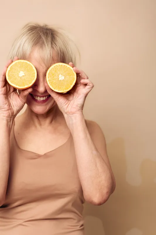 Happy middle aged woman with short blonde hair holds two orange halves in front of eyes, smiling in beige top. Concept of healthy lifestyle, vitamin C, fun and natural wellness.